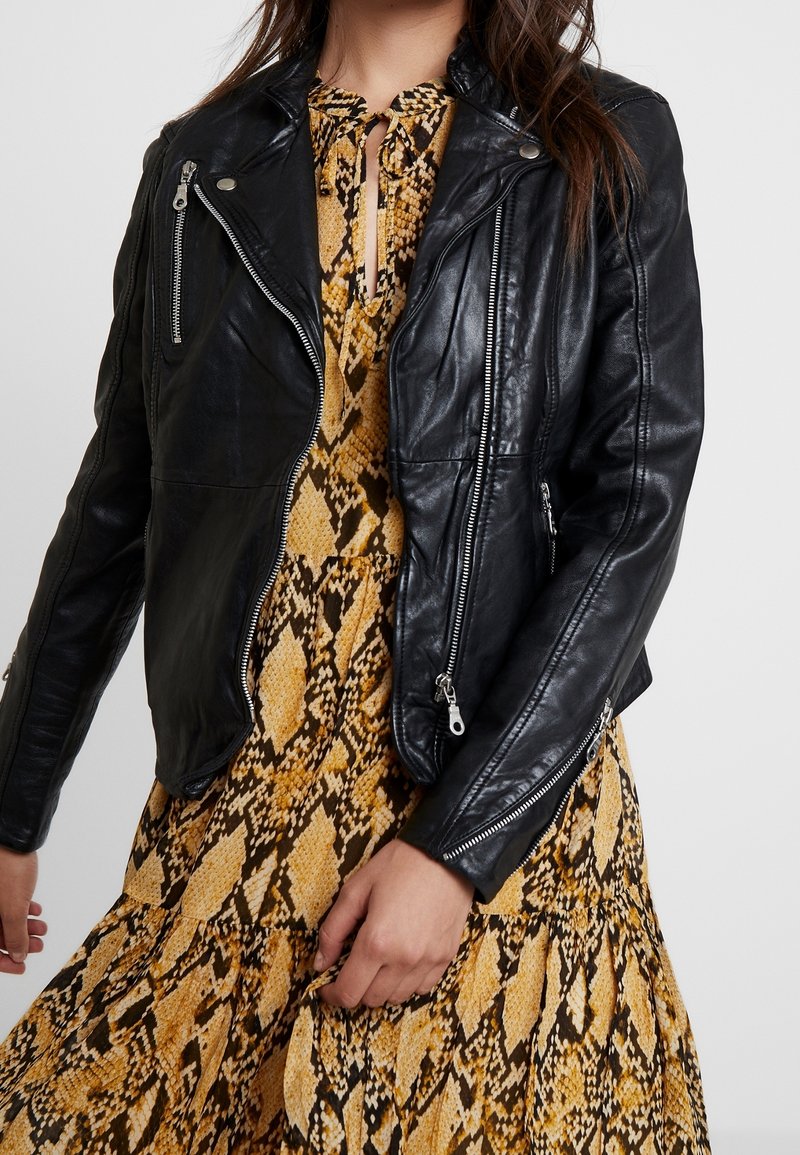 Giacca in pelle nera da motociclista con zip argentate, design aderente, davanti asimmetrico e finitura texturizzata, indossata sopra un vestito giallo con stampa a serpente.