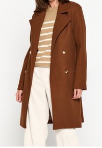 Vrouw met een bruine double-breasted jas, beige gestreepte trui en witte broek, staand tegen een effen achtergrond.