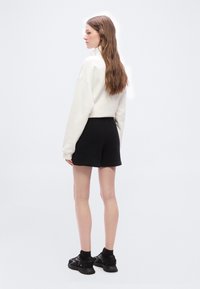 Γυναίκα με μακριά καστανά μαλλιά, ντυμένη με λευκό φούτερ, μαύρα σορτς και μαύρα αθλητικά παπούτσια, όρθια και κοιτάζοντας πλάγια σε λευκό φόντο.