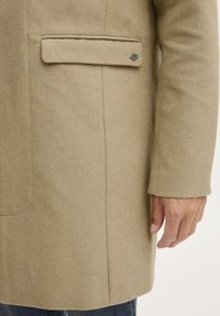 Cappotto di lana beige con una tasca sul petto, colletto elegante e linee pulite, realizzato in tessuto texturizzato e con dettagli metallici sottili.