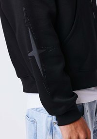 Sweat-shirt noir à manches longues avec un motif en forme d'étoile sombre sur la manche, porté avec un jean bleu clair et une chemise blanche en dessous.