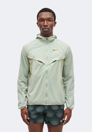 Homme portant une veste à capuche zippée vert clair et un short noir à motifs, debout devant un fond clair uni.