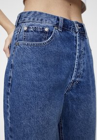 Jean en denim bleu avec taille haute, design classique à cinq poches, usures légères et détails en métal.