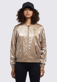 Metalická béžová bomber bunda s žebrovanými manžetami a lemem, obsahující přední zip a boční kapsy. Černá kšiltovka dokončuje vzhled.