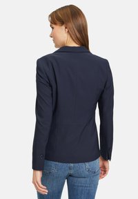 Námořnický modrý blazer s přiléhavým střihem, strukturálními rameny a detalem jednoho knoflíku na manžetě. Hladká textura s čistým designem zad.