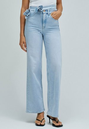 Femme portant un jean large taille haute bleu clair avec un détail décoratif en forme de rose à la taille, associé à des sandales à talons noires et un top blanc.