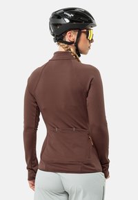 Maglia da ciclismo a maniche lunghe marrone con colletto alto, tasche posteriori e tessuto elastico per un comfort ottimale nei movimenti. Indossata con un casco nero e occhiali da sole.