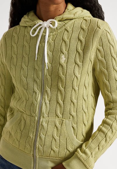 Asmuo dėvi šviesiai žalią megztą užsegamą su užtrauktuku gobtuvą su priekinėmis kišenėmis ir baltomis raištelėmis, surištomis kaspinėliu.