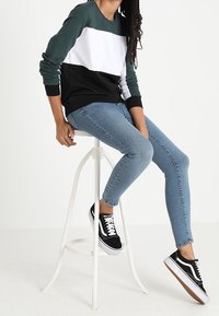 Felpa color block nera, bianca e verde, in misto cotone, con polsini a coste, abbinata a jeans slim-fit azzurri e scarpe nere stringate.