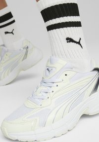 Baskets blanches en tissu aéré et panneaux synthétiques, accents réfléchissants argentés, semelle épaisse, accompagnées de chaussettes rayées noir et blanc.