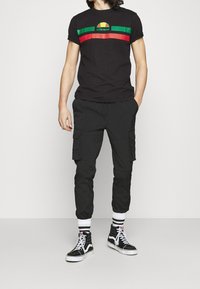 Camiseta negra con un logotipo colorido y diseño a rayas, combinada con pantalones cargo negros que cuentan con bolsillos laterales y zapatillas altas negras.