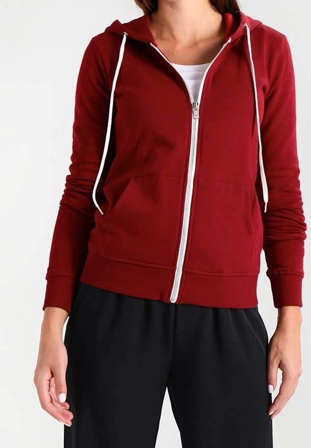 Femme portant un sweat à capuche rouge zippé avec des cordons blancs, un haut blanc et un pantalon noir ample sur un fond uni.