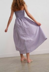 Abito maxi in gingham lilla con corpino arricciato. Tessuto fluido e silhouette dritta. Completato da semplici sandali infradito.