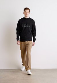 Zwarte sweatshirt met grijs 'BOSS' logo, beige tapered pants en witte sneakers; model loopt tegen een eenvoudige achtergrond.