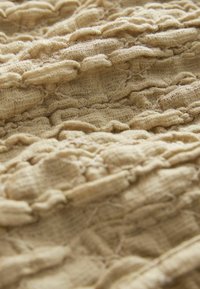 Texturiertes, hellbraunes Gewebe mit erhabenen, gewellten Mustern und einer weichen, gestrickten Optik. Zeigt ausgefeilte Details und eine leicht gekräuselte Oberfläche.