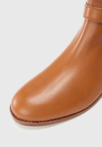 Primer plano de una bota de tobillo suave y marrón claro con punta redondeada y un sutil detalle de costura. El fondo es blanco liso.