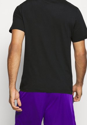 Personne vue de dos portant un t-shirt noir à manches courtes et un short de sport violet, debout avec les bras détendus le long du corps.