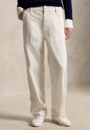 Άτομο που φοράει μπεζ παντελόνι με ίσια μπατζάκια και μεγάλες εμπρόσθιες τσέπες, σε συνδυασμό με λευκά δερμάτινα παπούτσια με κορδόνια και μπλε πουλόβερ με γυρισμένα μανίκια.