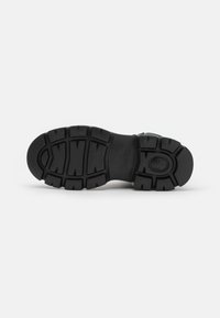 Suela de zapato de goma negra con un patrón de banda de rodadura grueso; presenta secciones elevadas y un logo cerca del talón para mayor tracción y durabilidad.