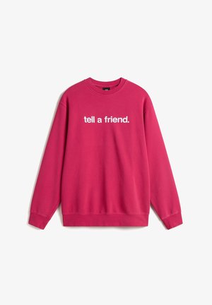 Personne portant un sweat-shirt rose avec un texte blanc partiellement visible et un pantalon marron foncé, debout devant un fond blanc uni.