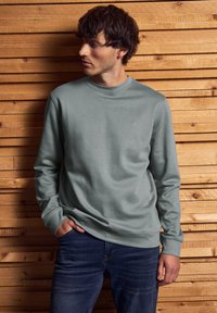 Muž v světle šedé mikině a tmavých džínech, stojící s jednou rukou v kapse před pozadím z na sebe naskládaných dřevěných prken.