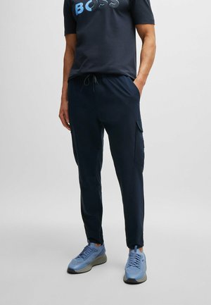 Mężczyzna ubrany w czarny t-shirt BOSS, czarne spodnie z troczkami i kieszeniami cargo oraz niebieskie sportowe buty, stoi na jednolitym tle.