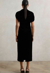 Čierne sametové šaty s krátkymi rukávmi, so zberaním na chrbte, dĺžka po kolená a jemná textúra. Skombinované s čiernymi sandálmi na remienky.
