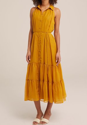 Femme portant une robe midi sans manches jaune moutarde avec des boutons sur le devant et une jupe à volants étagée, associée à des sandales blanches à enfiler.