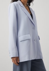 Světle modrý blazer z hladké tkaniny, s klopovými límci, dvěma předními kapsami a zapínáním na jeden knoflík. Dlouhé rukávy s knoflíkovými doplňky.