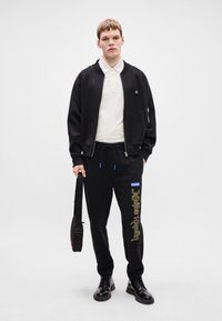 Μαύρο μπουφάν bomber με φερμουάρ πάνω από λευκό πόλο, σε συνδυασμό με μαύρους τζόγκινγκ με κίτρινο κείμενο και λογότυπο. Μαύρα παπούτσια και μαύρη τσάντα.