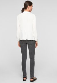 Witte geplooide blouse met lange mouwen en een gerimpelde halslijn, gecombineerd met donkergrijze skinny jeans en zwarte platte sandaaltjes.