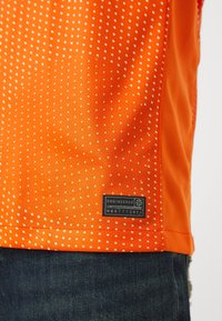 Chemise courte à manches courtes de couleur orange vif avec une texture en pointillés. Présente une étiquette noire avec du texte blanc à l'ourlet. Jean en denim bleu visible en dessous.