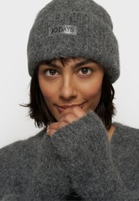 Gorro de lana gris con una superficie texturizada y un parche con logo. El modelo lleva un suéter gris de gran tamaño con puños anchos que hace juego.