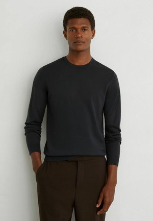 Mand står mod en hvid væg iført en tætsiddende sort langærmet sweater og mørkebrune bukser med den ene hånd i lommen.