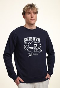 Sudadera azul marino con un diseño gráfico blanco que incluye el texto "SHIBUYA 1961 Tokio" y un personaje de béisbol. Mangas largas y cuello redondo.
