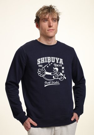 Marineblauwe sweatshirt met een witte grafische opdruk met de tekst "SHIBUYA 1961 Tokyo" en een honkbalpersonage. Lange mouwen en ronde hals.