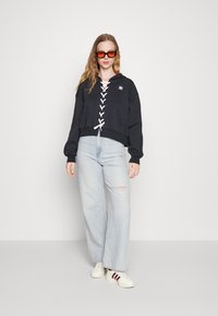 Cappuccio nero corto con dettagli in pizzo bianco, abbinato a jeans a gamba larga di un lavaggio chiaro e sneakers bianche con strisce rosse.