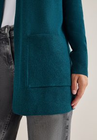 Tyrkysový pletený kardigan s texturovaným povrchem, žebrovanými manžetami a lemem. Obsahuje praktickou náprsní kapsu.
