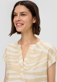 Beige Bluse mit weißen Zebra-Streifen, kurzen Ärmeln, V-Ausschnitt und weicher Textur, mit entspannter Passform und dezenten Designdetails.