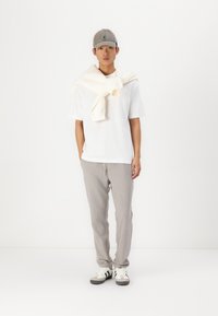 Weißes T-Shirt, hellgraue Jogginghose, weiße Turnschuhe mit schwarzen Streifen und graue Mütze. Beige Pullover über die Schultern drapiert.