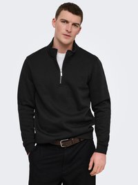 Jeune homme portant un pull noir à fermeture éclair quart, un sous-vêtement blanc, un pantalon noir et une ceinture marron, debout devant un fond clair uni.