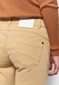 Primer plano de una persona que lleva pantalones beige con un bolsillo trasero visible y una etiqueta blanca, combinados con una camiseta de manga larga marrón de canalé.