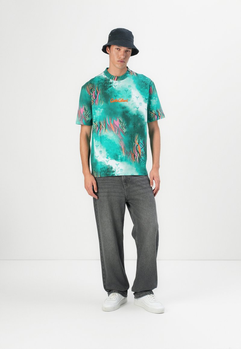 Grünes T-Shirt mit kurzen Ärmeln, buntem tropischen Druck und gesticktem Text, kombiniert mit lockeren grauen Jeans aus Denim und weißen Sneakers.