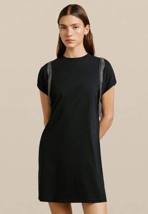 Mujer vistiendo un sencillo vestido negro de manga corta con detalles de flecos de cadena plateada en los hombros, de pie frente a un fondo beige liso.