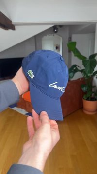 Ruka drží modrú čiapku Lacoste s bielym písmom a zeleným logom krokodíla v obývacej izbe s drevenou podlahou a kvetináčom.