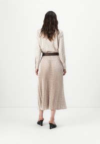 Jupe longue plissée en tissu à motifs beiges clairs, complétée par une ceinture marron foncé. Le modèle porte des mules à talons noirs.