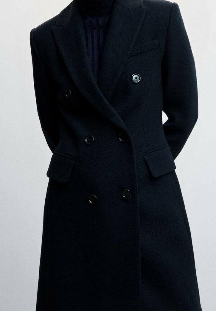 Manteau en laine noir à double boutonnage avec revers crantés, deux poches à rabat et boutons noirs, porté sur un haut foncé à col montant.