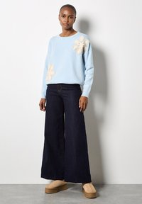 Suéter de punto azul claro con grandes aplicaciones de flores color crema. Combinado con pantalones de mezclilla oscuros de pierna ancha y zapatos de plataforma beige. Textura suave.