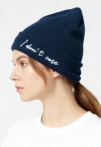 Cappellino lavorato a maglia blu navy con risvolto. Presenta un ricamo bianco con la scritta "Non mi importa." Texture liscia con vestibilità aderente.