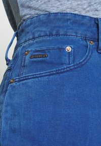Blå denimjeans med slim fit. Funktioner inkluderar en framficka med synlig sömnad, märkesmetalldetaljer och texturerad tygliknande detaljer.